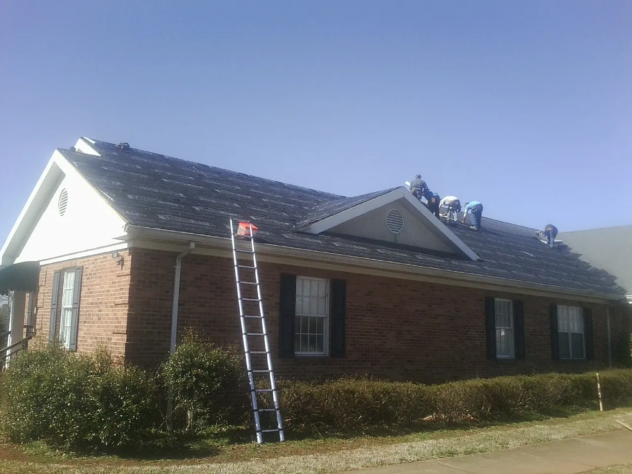 Crew tearing shingles off a brick ranch roof with a ladder set — Metal Roof Repair work by our crew in Phillips