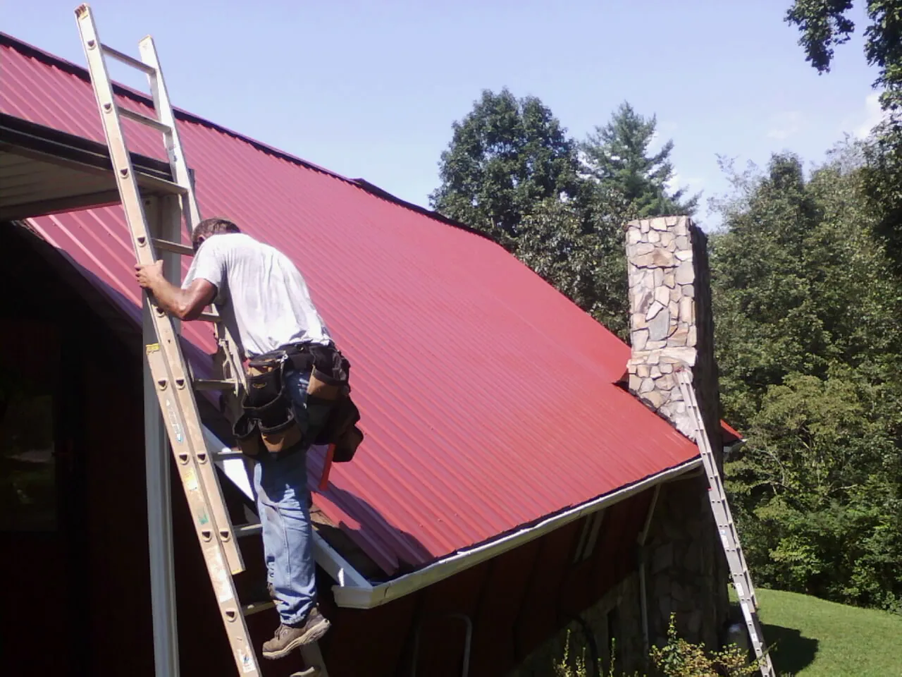 Roofer on a ladder beside a red metal roof and stone chimney — Soffit & Fascia Repair by Roofing Brothers in Phillips